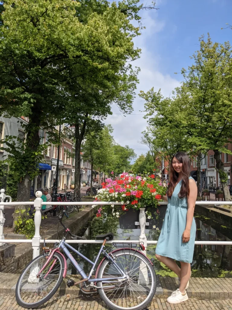 Travel blog author Sophie Chen standing on a white canal bridge next to a bicycle and vibrant flower boxes in a scenic canal street in Amsterdam